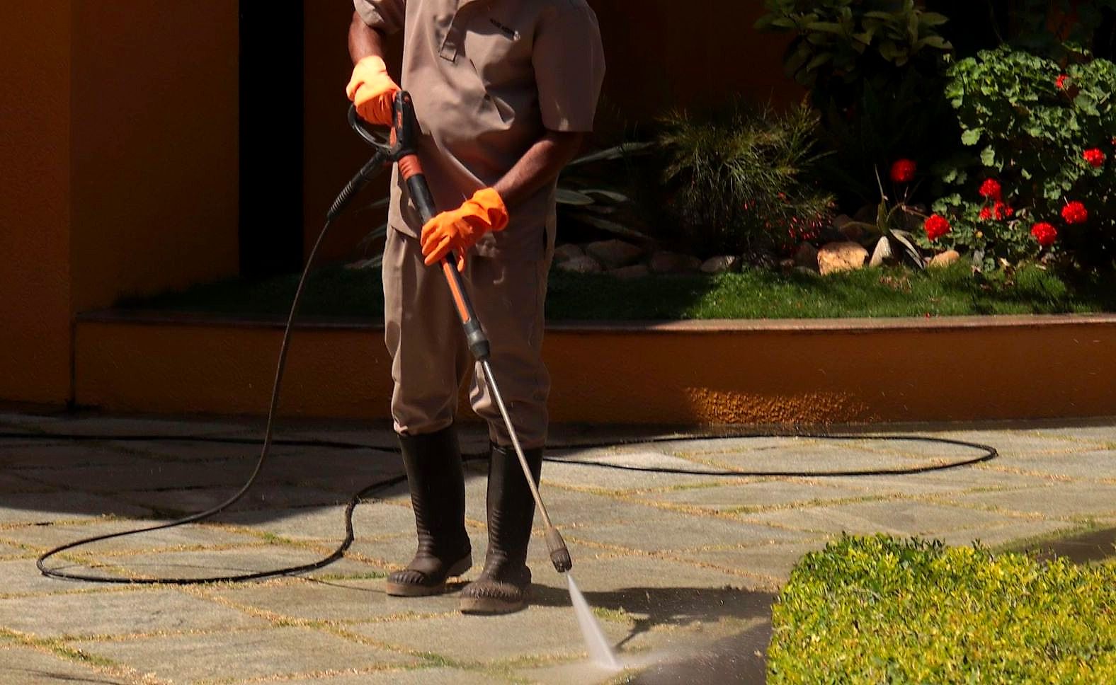 A gardener wearing a hat cleans an urban sidewalk with a pressure washer.